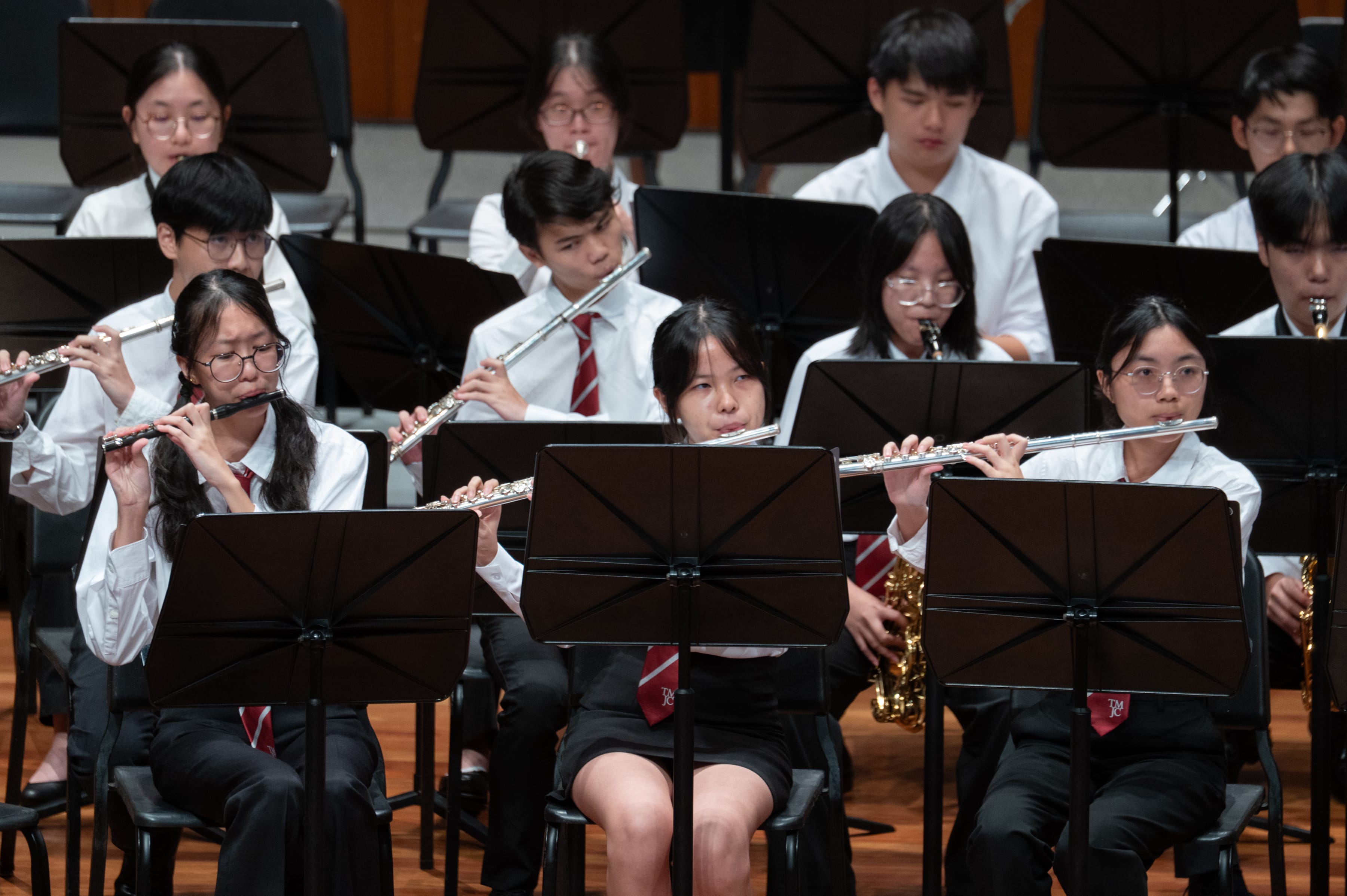 A close-up of the woodwind section of a student symphonic band in performance, with several students in white shirts and red ties playing silver flutes and a saxophone, reading from music stands in a concert hall setting.